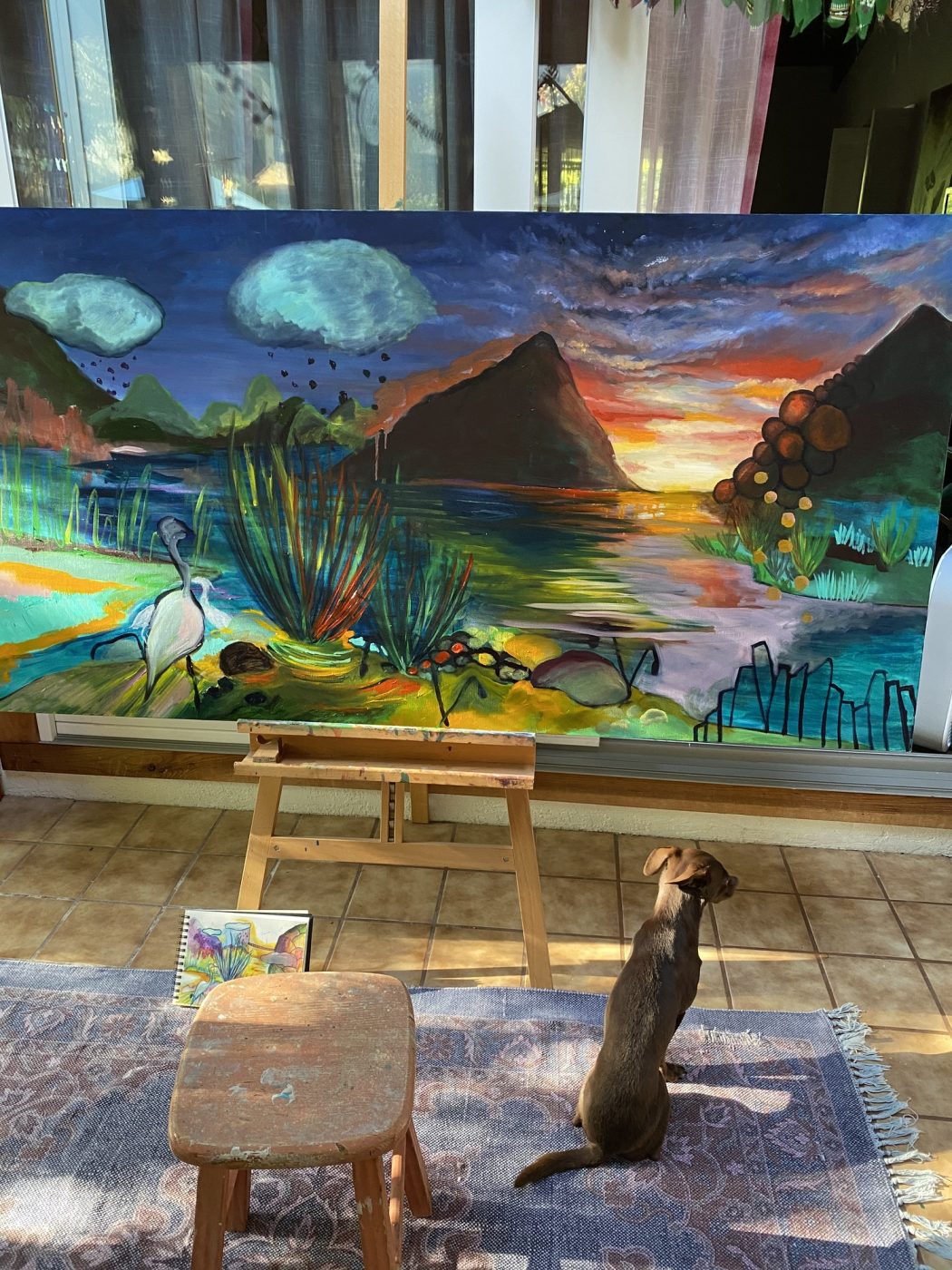 A landscape painting on an easel in the studio, with Amy’s dog sitting beside it.