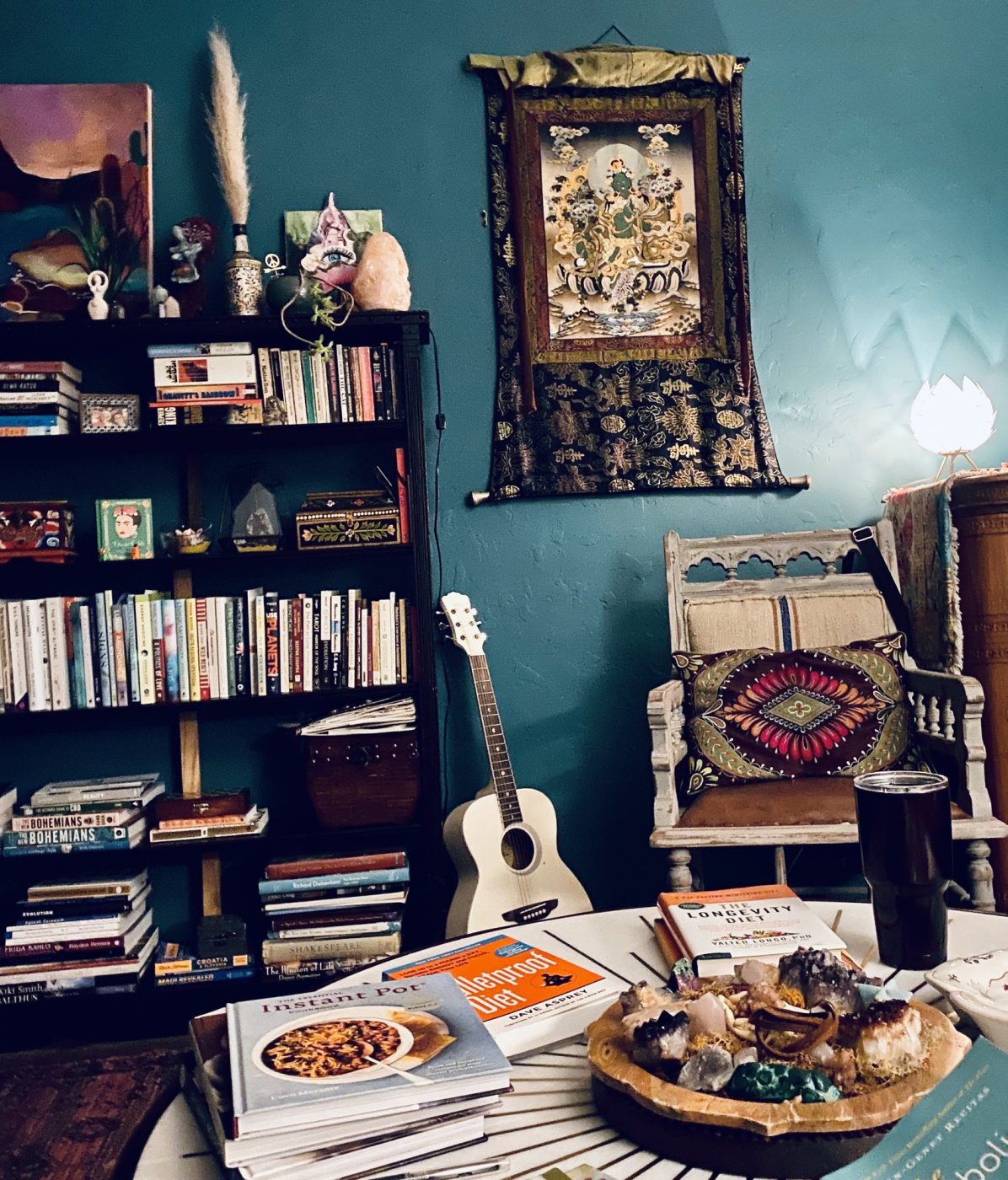 A teal wall with bookshelves, a guitar and a painted thangka above a vintage chair.